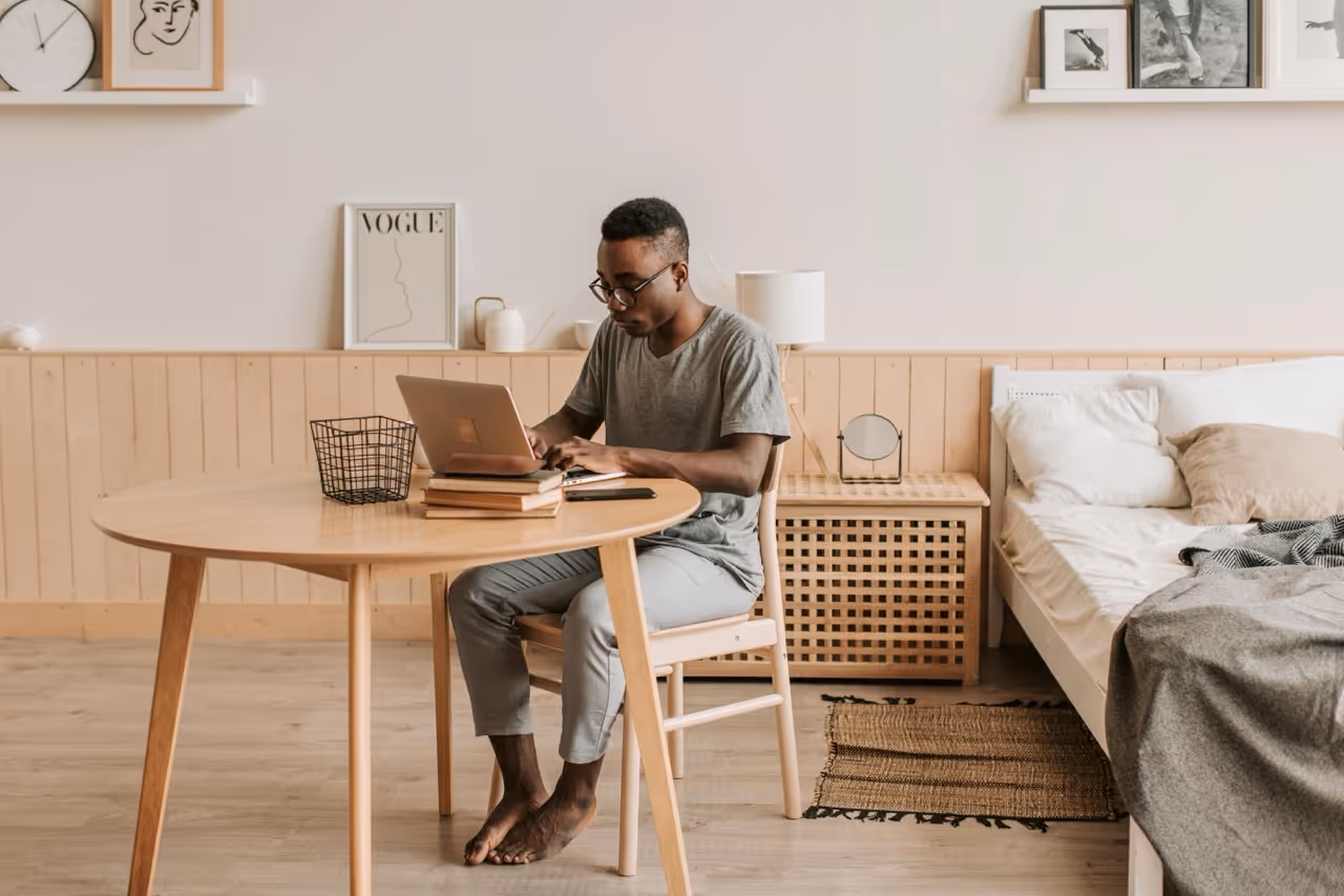 Man wearing glasses sitting barefoot at a wooden table working on a laptop in a minimalist bedroom.