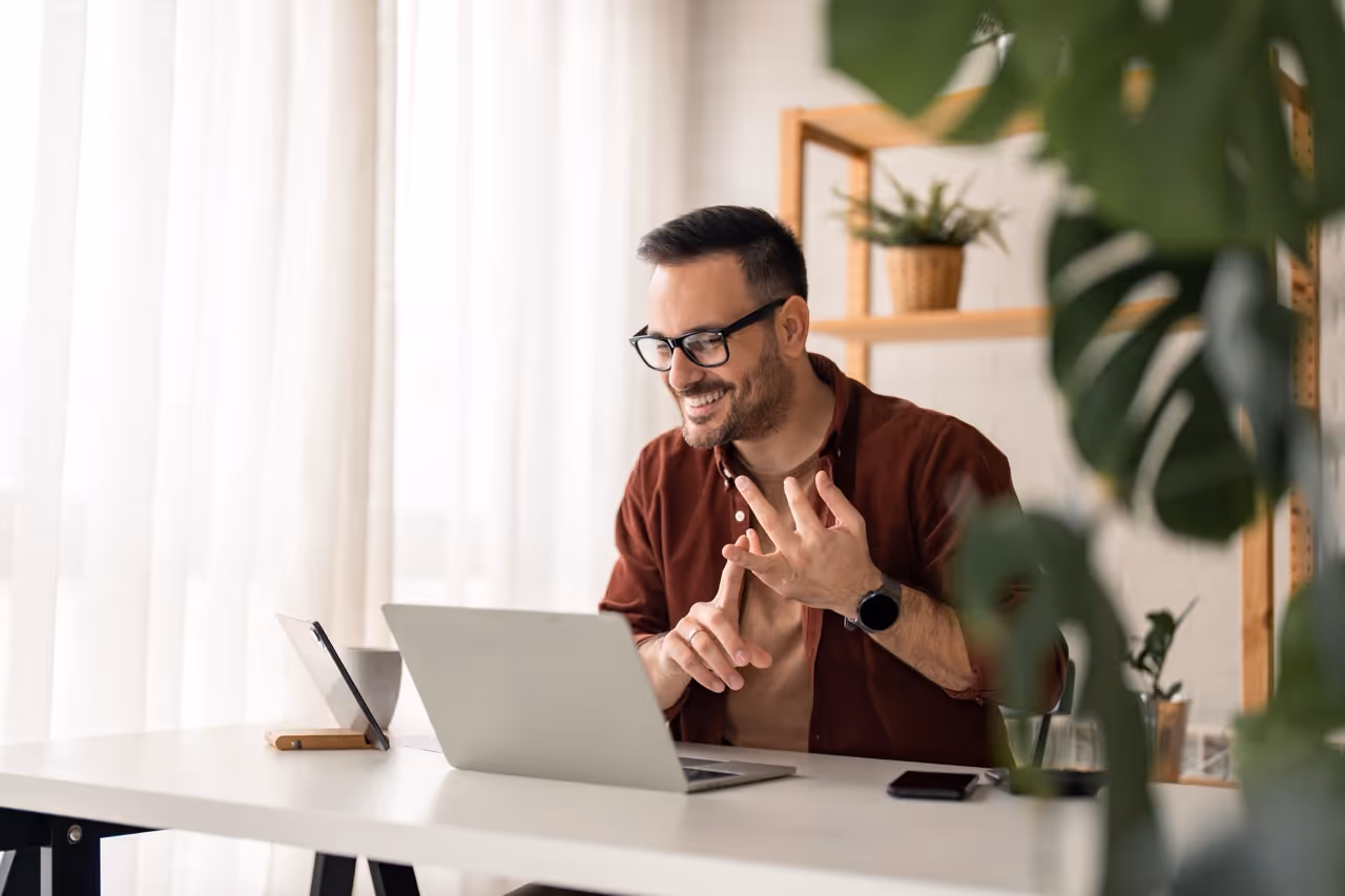 Smiling man wearing glasses counting on his fingers while sitting at a desk with a laptop in a bright room.