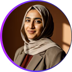 Lächelnde Frau mit einem hellbraunen Hijab und einem braunen Blazer vor einem unscharfen, neutralen Hintergrund.