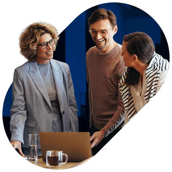 Three colleagues smiling and discussing work around a laptop in an office setting.