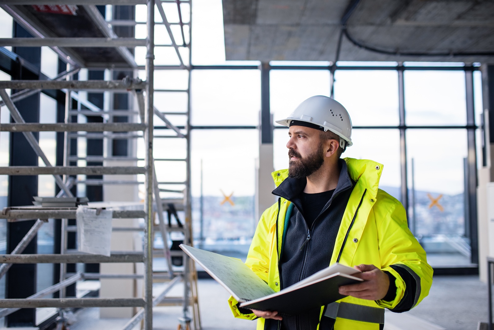 Bearded manager in yellow safety jacket and white hard hat holding a folder inside a construction site.
