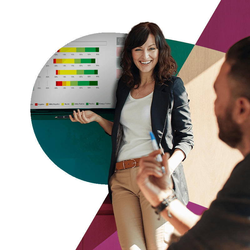 A woman in business casual attire smiling and pointing at a presentation board with colorful horizontal bar charts during a meeting with a man holding a marker.
