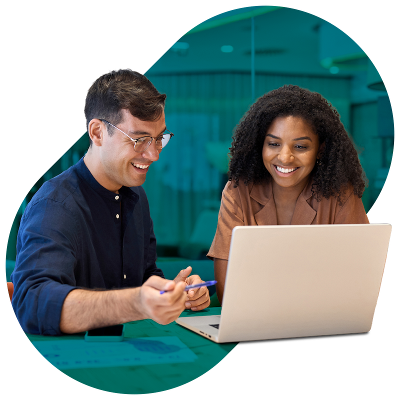 A man and woman sitting together at a table, smiling and looking at a laptop screen, with the man holding a pen and pointing at the screen.