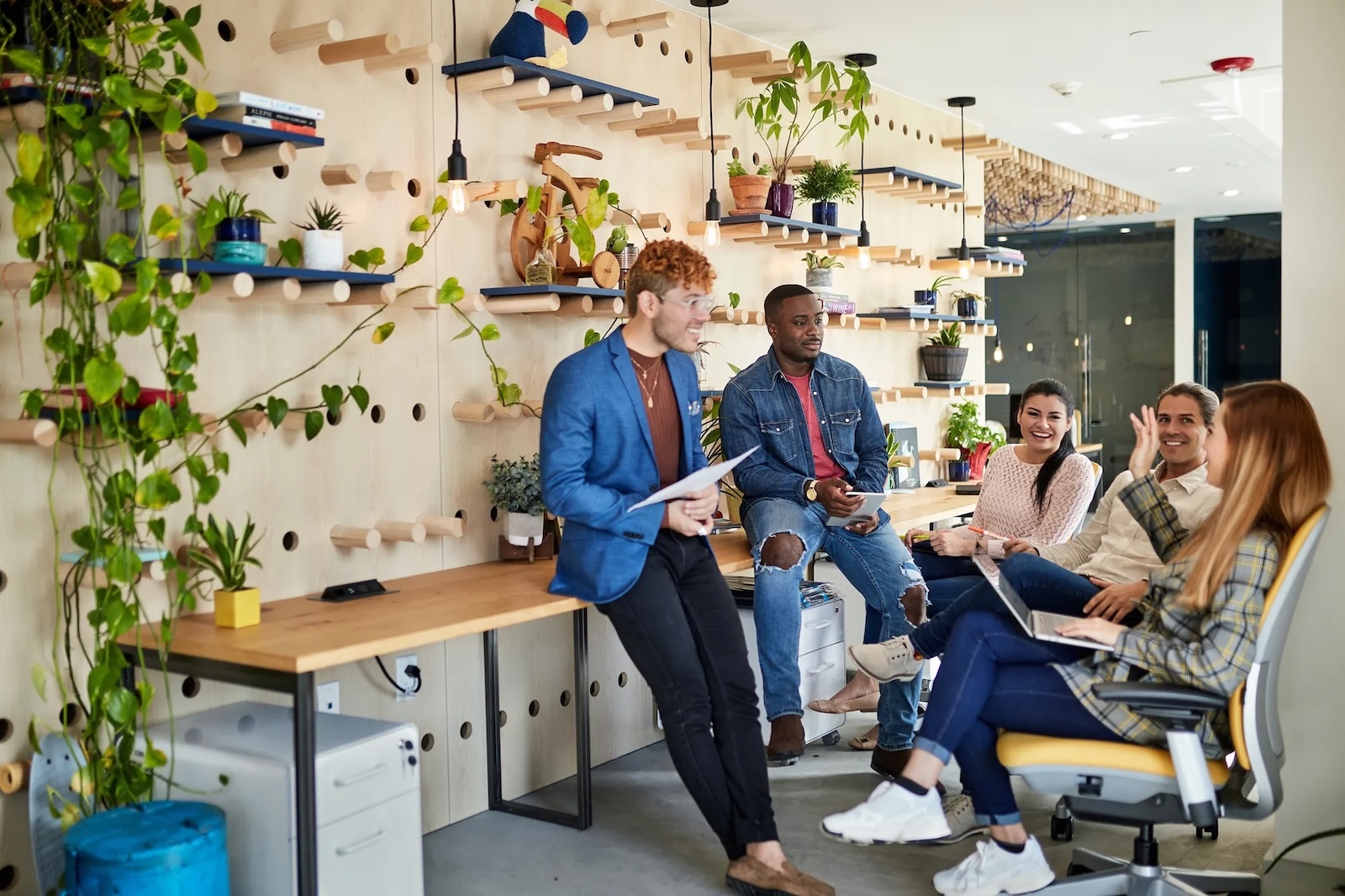 Five young professionals casually chatting in a modern office with plants and wooden shelves.