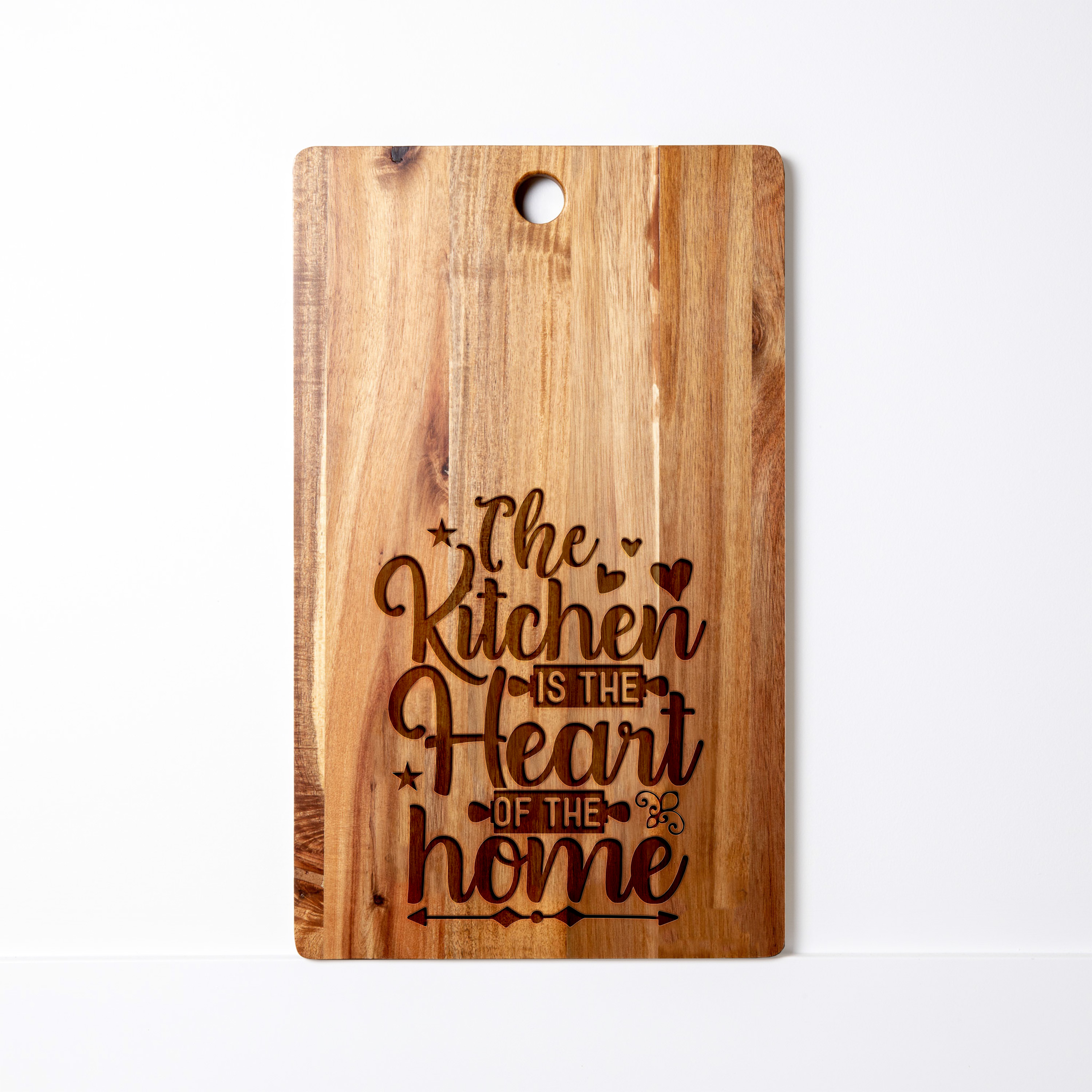 Acacia wood cutting board engraved with the phrase ‘The Kitchen is the Heart of the Home’ in decorative script with heart and star accents.