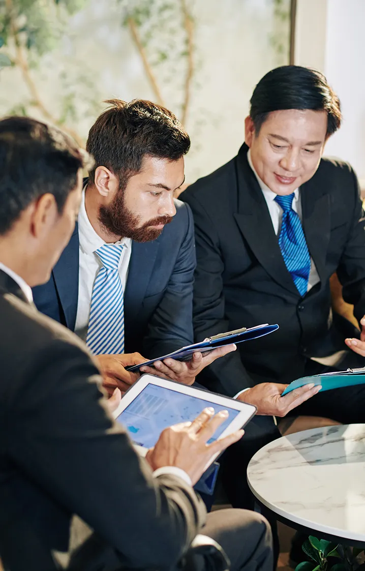 Three business professionals in suits discussing documents and a tablet around a small round table.