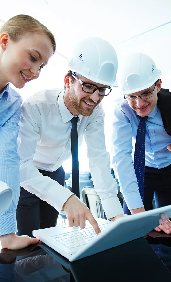 Three professionals in business attire with white hard hats looking at a laptop on a table in a bright office.
