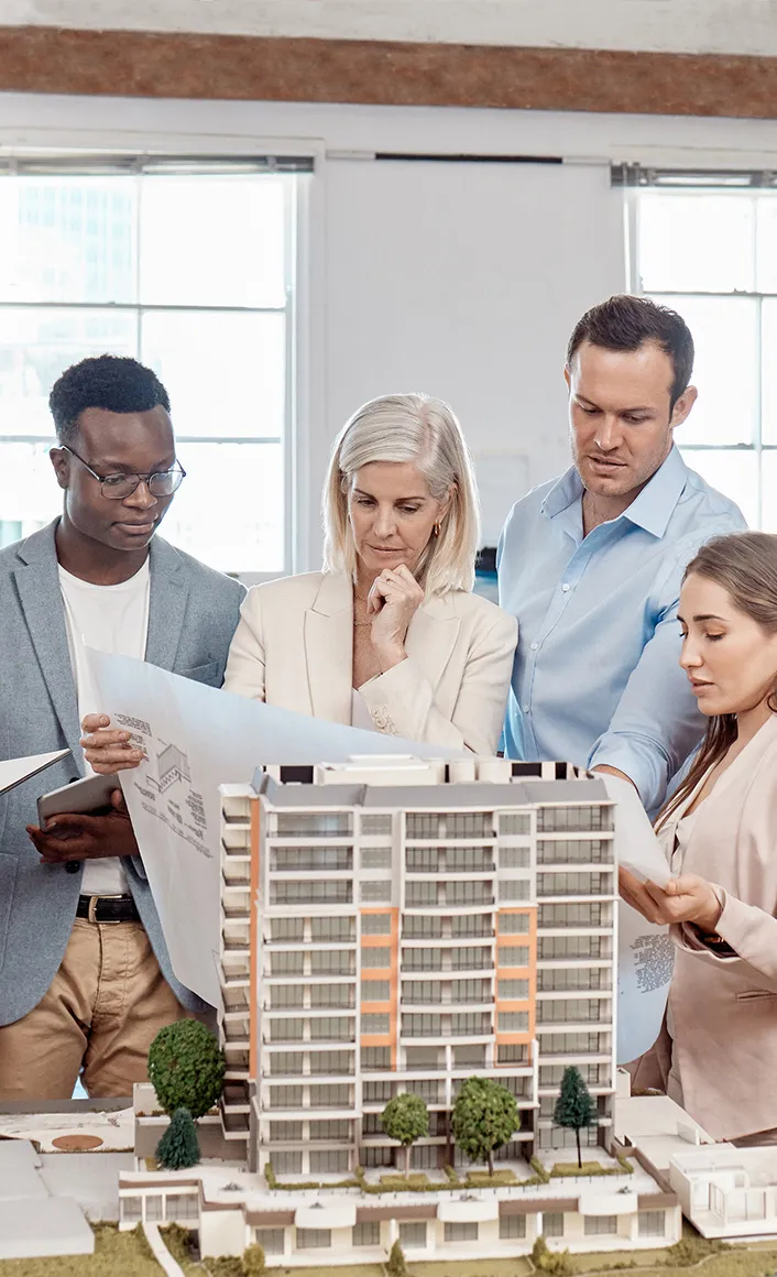Five diverse professionals reviewing architectural plans around a detailed scale model of a modern multi-story building in a bright office.