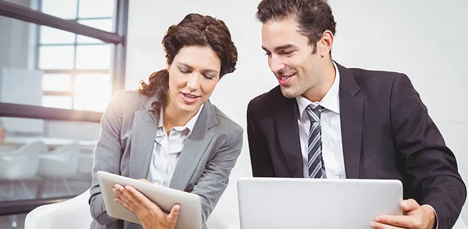 A businesswoman showing a tablet to a businessman who is holding a laptop in a modern office.