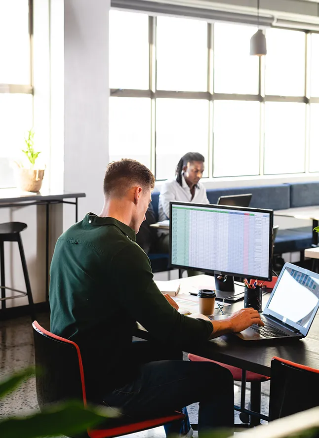 Man working at a desk with a laptop and a large monitor displaying a spreadsheet in a bright office.