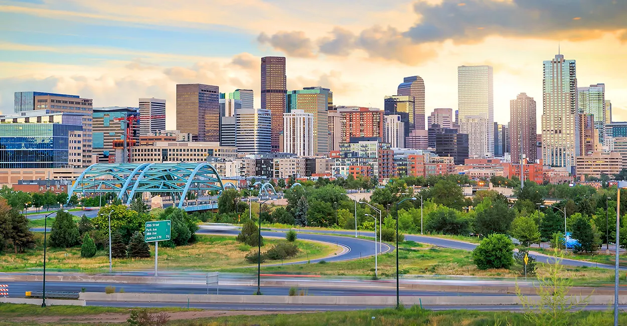 Denver city skyline at sunset with modern buildings, a blue arched bridge, and curving roads surrounded by greenery.