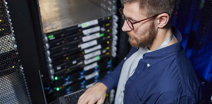 Technician wearing glasses working on a laptop in front of server racks in a data center.