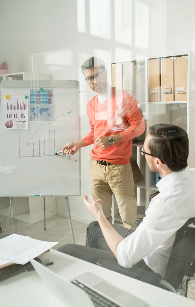 Man in orange sweater presenting bar chart on whiteboard to seated colleague in office.