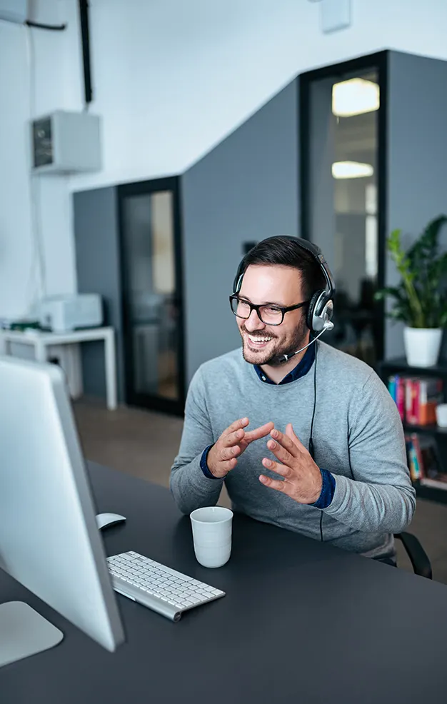 Smiling man wearing glasses and headset talking on a video call at his desk in a modern office.
