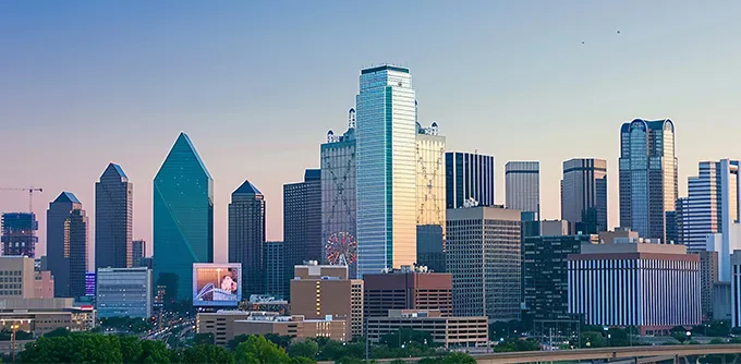 Dallas city skyline at sunset with modern skyscrapers and green trees in foreground.