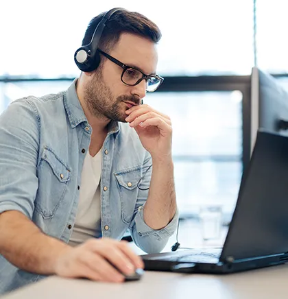 Man wearing glasses and headphones working on a laptop at a desk, looking focused.