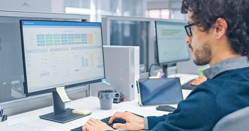 Man wearing glasses working on a computer displaying a project management dashboard in an office.