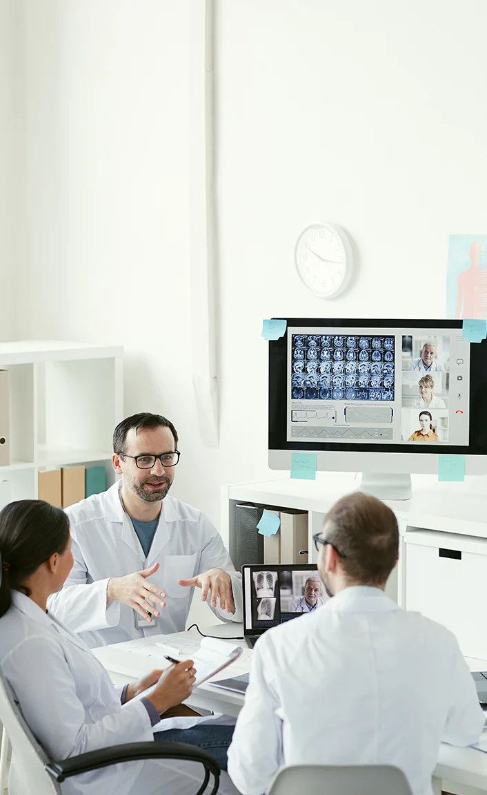 Three doctors in white coats discussing X-ray and brain scan images during a video conference in a bright office.