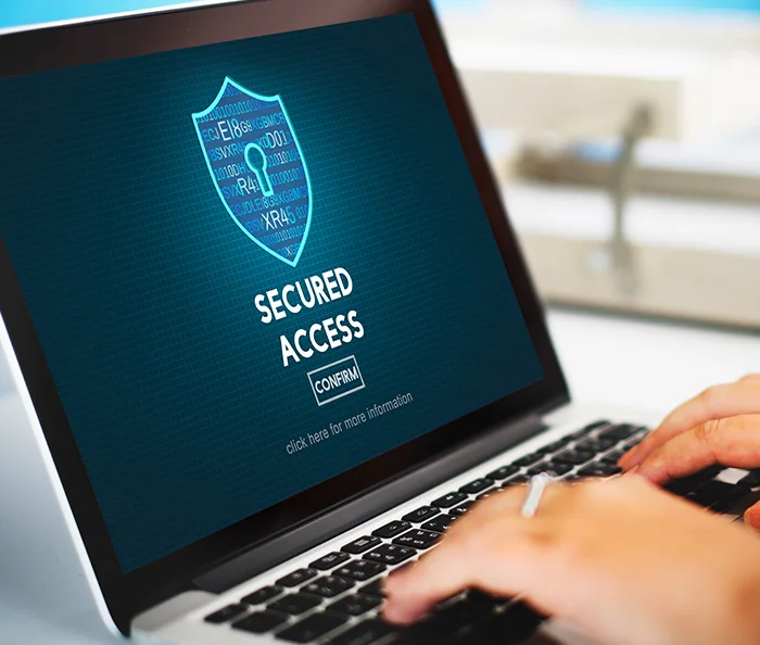 Laptop screen showing a shield with a keyhole and text reading 'SECURED ACCESS' with a 'CONFIRM' button, hands typing on keyboard.