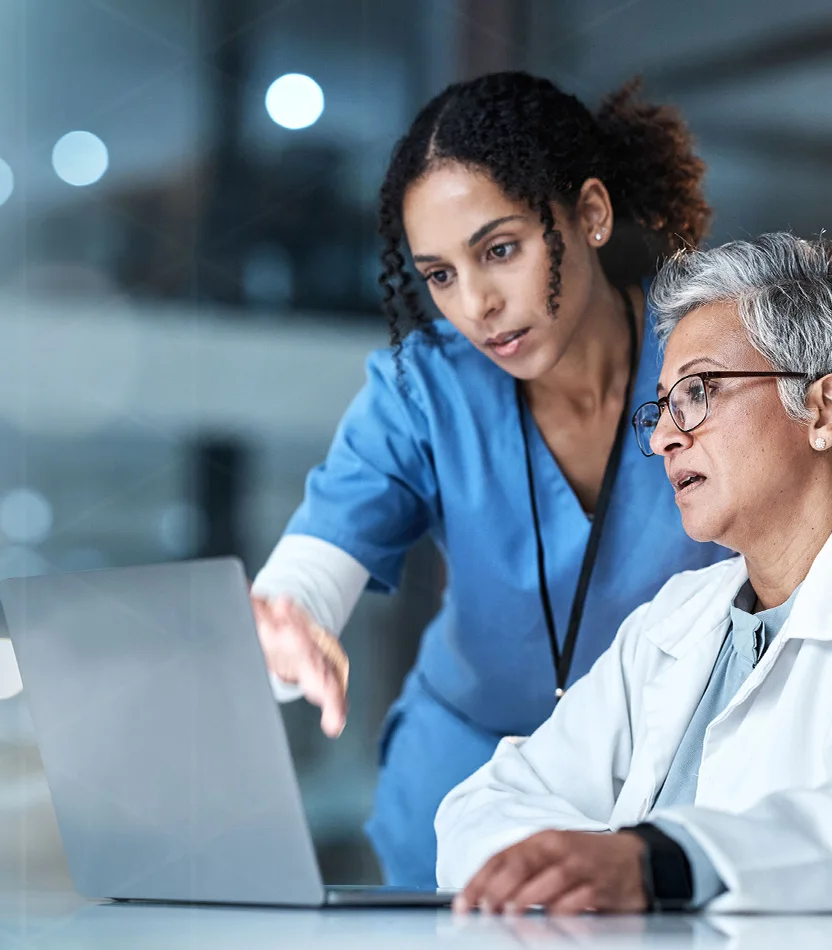 Two healthcare professionals, one in blue scrubs and one in a white coat, collaborating and looking at a laptop screen.