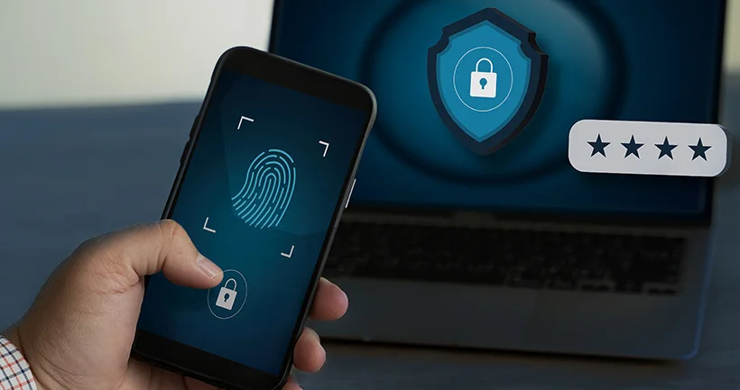 Hand holding smartphone displaying fingerprint scan and lock icon with a laptop showing a shield lock and four-star rating in the background.