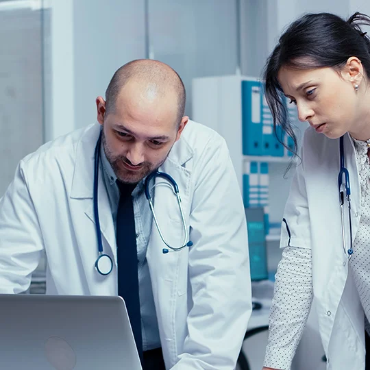 Two doctors in white coats with stethoscopes discussing something while looking at a laptop screen.