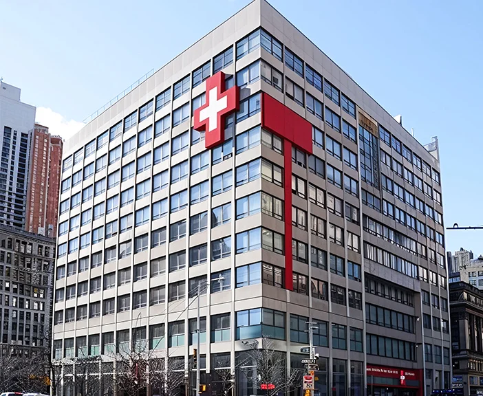 Modern multi-story hospital building with large red medical cross symbols on the facade, surrounded by city buildings under a clear sky.