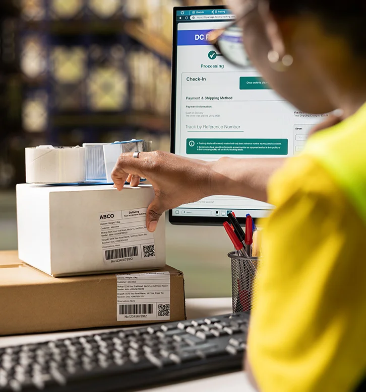 Person in yellow shirt managing shipping labels on packages with tracking information displayed on a computer screen.