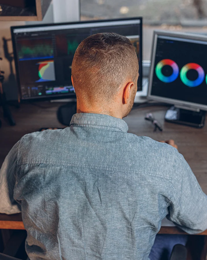 Man sitting at a desk working on video color grading with dual monitors displaying color wheels and editing software.