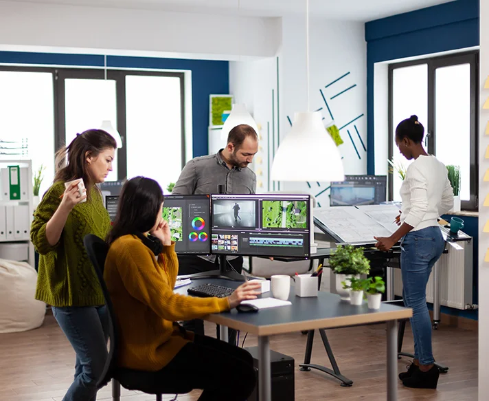 Four colleagues collaborating in a modern office, two women seated at a desk with dual monitors displaying video editing software, while a man and another woman review large printed plans nearby.