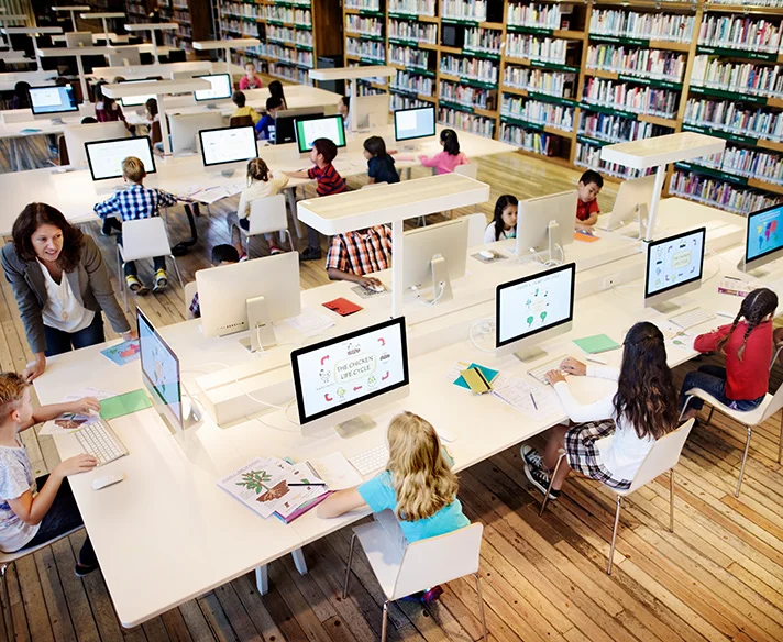 Children of diverse backgrounds working on computers in a school library with bookshelves and a teacher assisting a student.