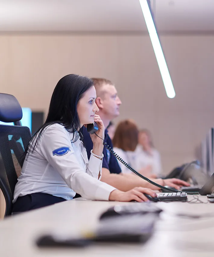 Female security guard speaking on a phone while working at a computer in a control room with other operators.