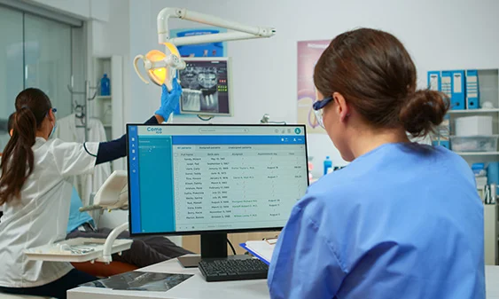 Orthodontist assistant in blue scrubs reviewing patient records on a computer screen while a dentist adjusts a dental light for a patient in a clinic.