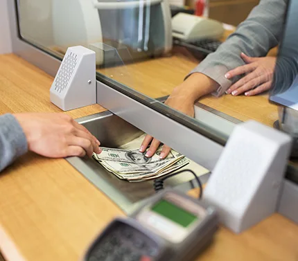 Person handing US dollar bills to a bank teller through a counter window with a payment terminal visible.