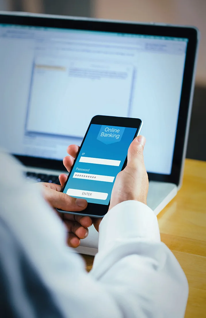 Person holding smartphone with online banking login screen showing password input and enter button, in front of a laptop.
