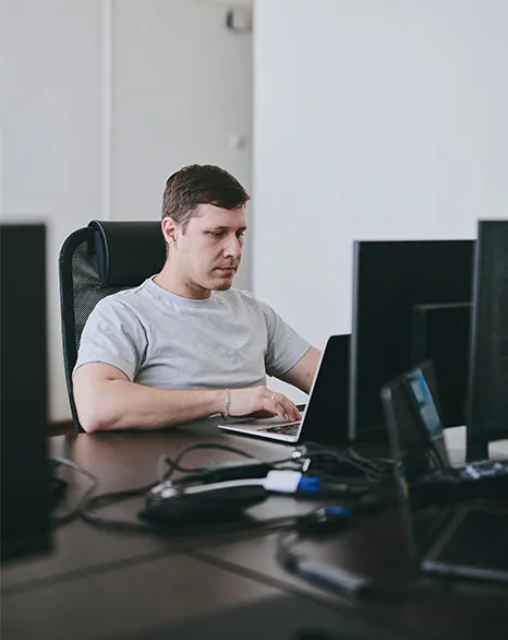 Man in a gray shirt working on a laptop at a desk with multiple monitors in an office.