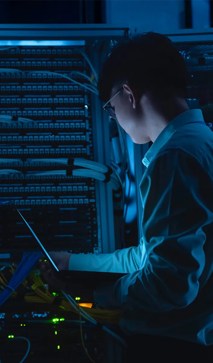 Technician working with server rack and cables in a dimly lit data center using a laptop.