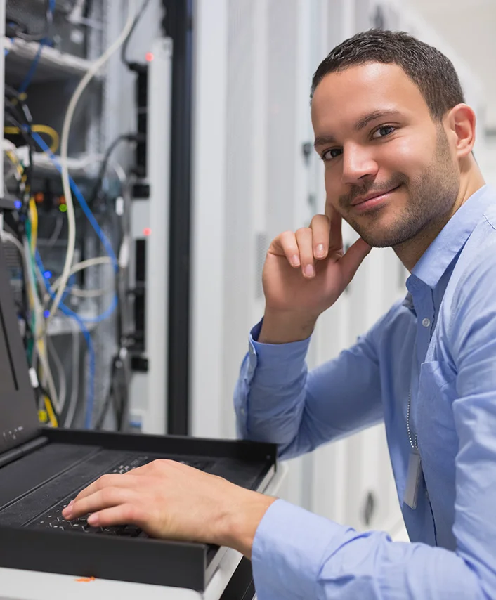 Man in blue shirt working on a laptop in a server room with networking cables and equipment.