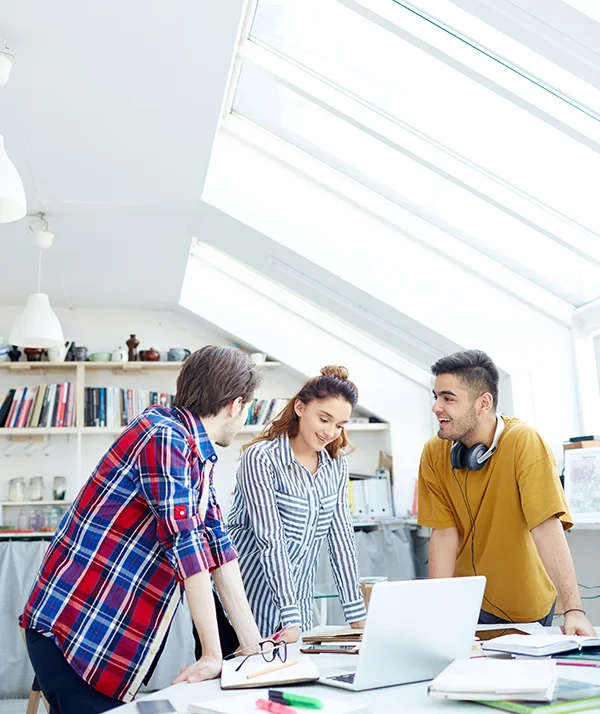 Three young coworkers collaborating at a desk with a laptop in a bright office with skylights.