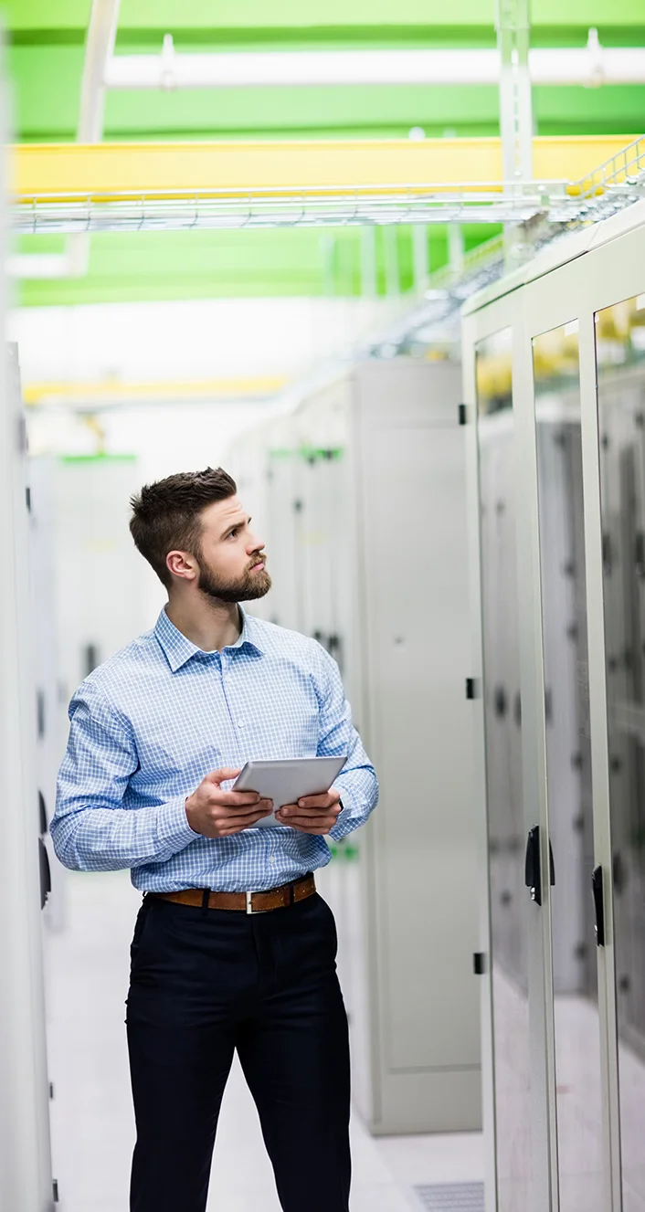 Man holding tablet inspecting servers in a data center with green and yellow ceiling pipes.