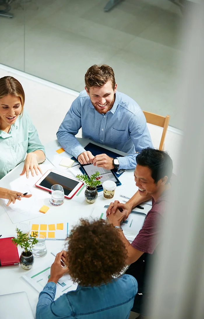 Four diverse colleagues smiling and discussing work around a white table with documents and devices.