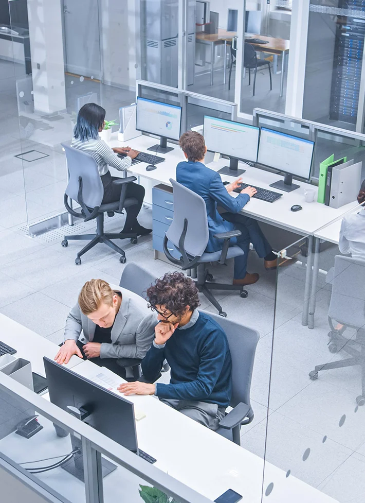 Four office workers focused on computers and documents inside a modern workspace with glass walls.