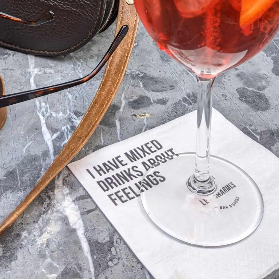 Wine glass with red beverage on a white napkin printed with the phrase 'I have mixed drinks about feelings' on a gray marble surface beside brown bag strap and sunglasses.