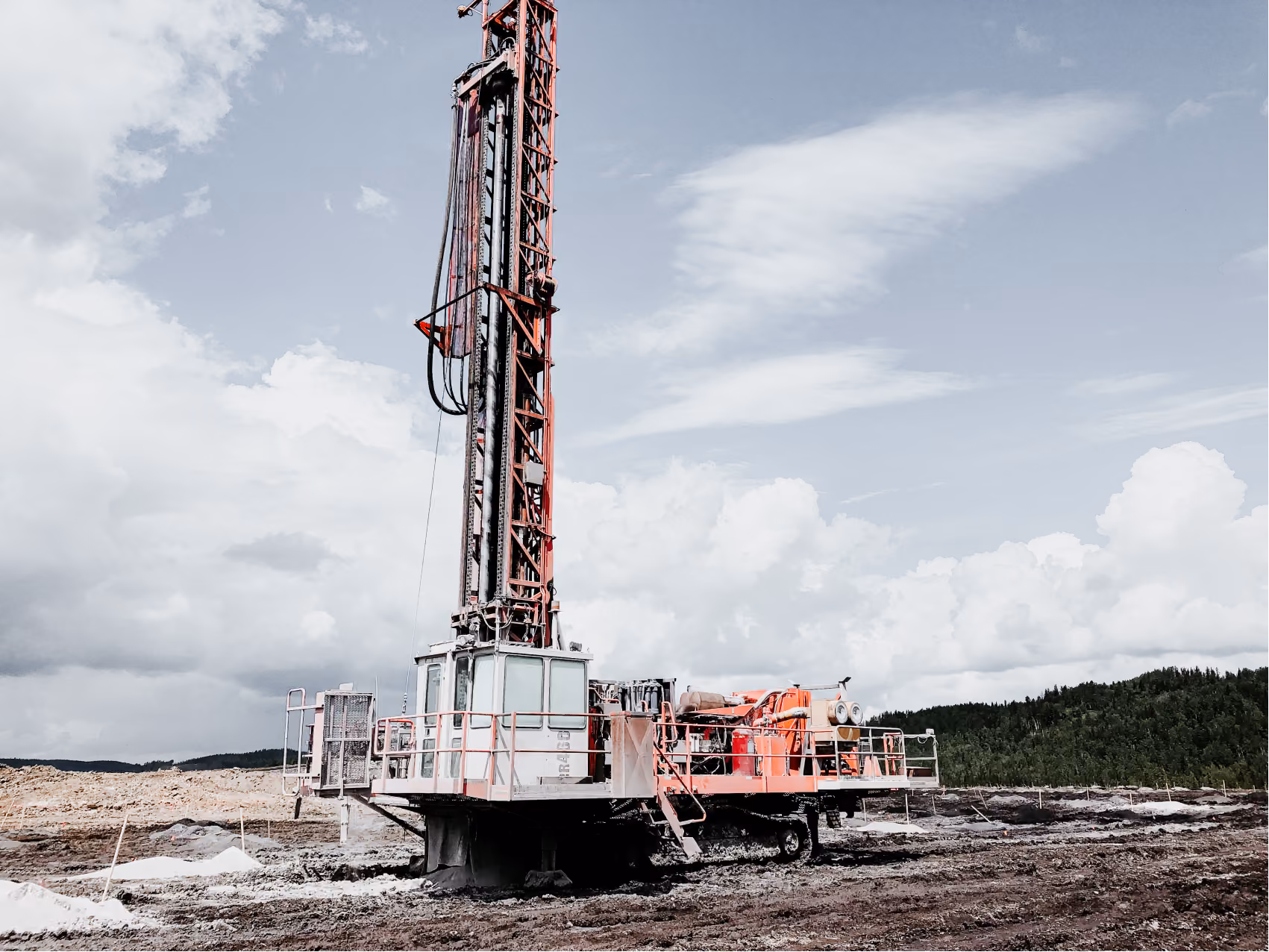 Large drilling machine on muddy ground in an open landscape with a cloudy sky and forested hills in the distance.