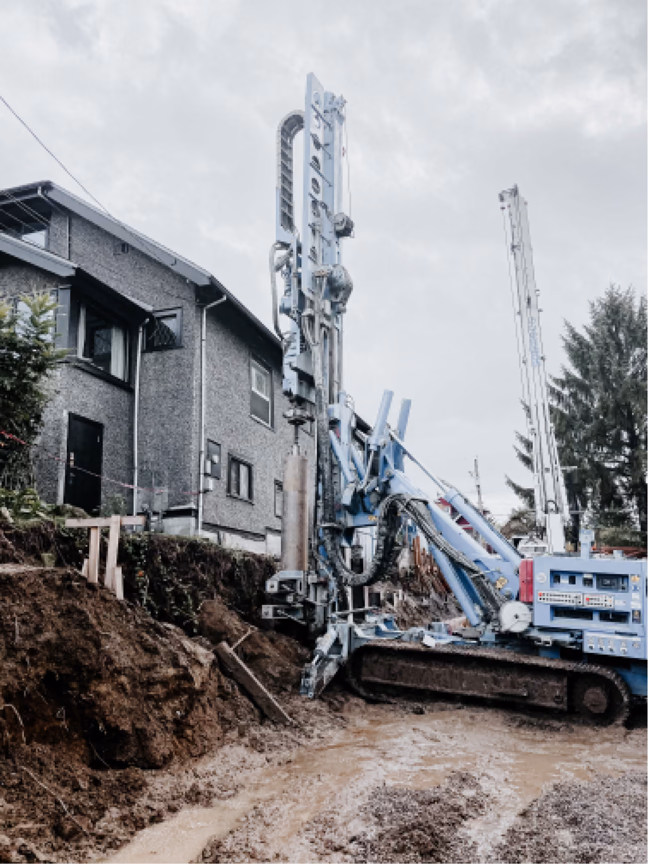 Large blue drilling machine with tracked base operating beside a residential building on a muddy construction site, excavating soil near a raised earth embankment.