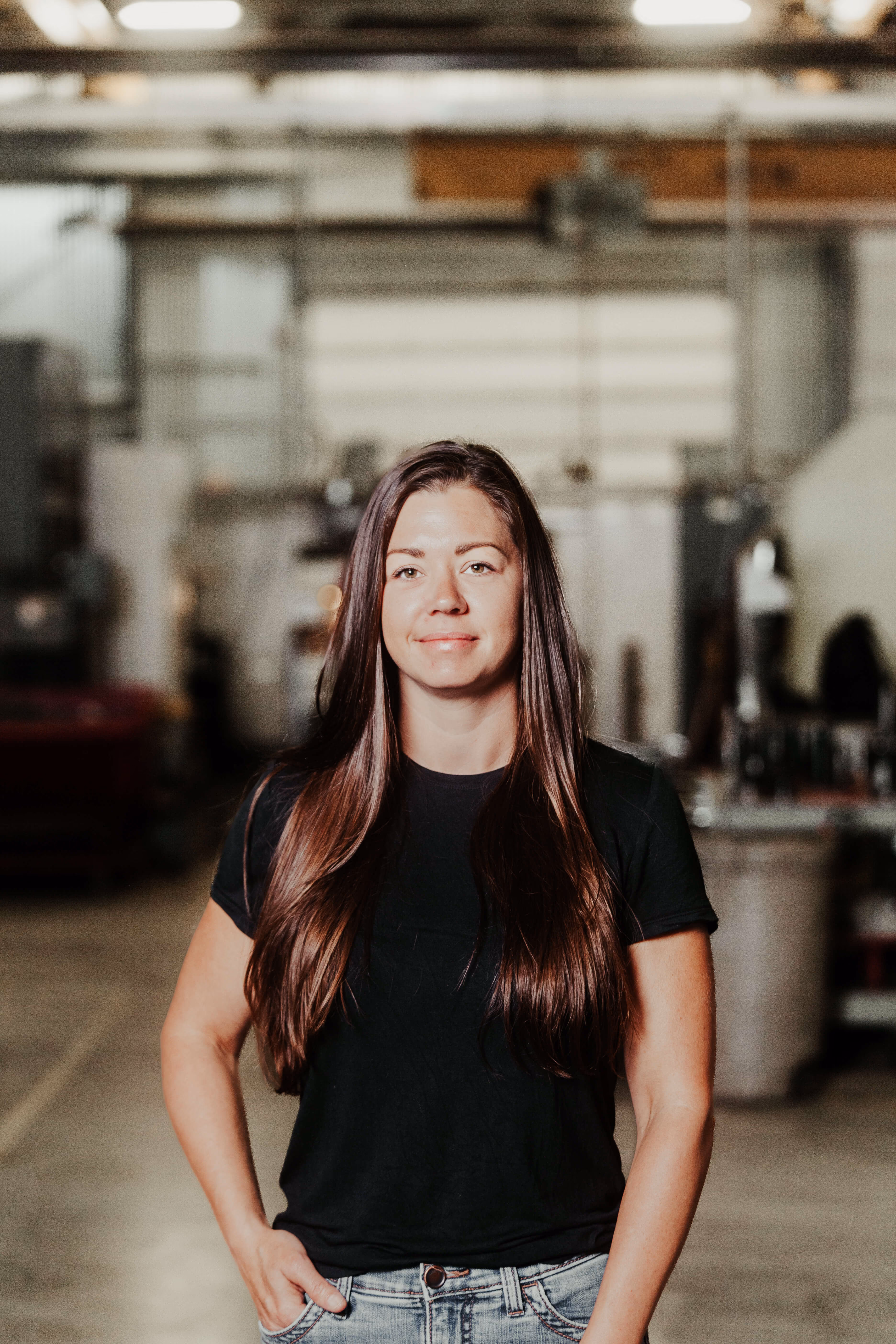 Woman with long brown hair wearing a black t-shirt and blue jeans stands with one hand in her pocket in an industrial setting.