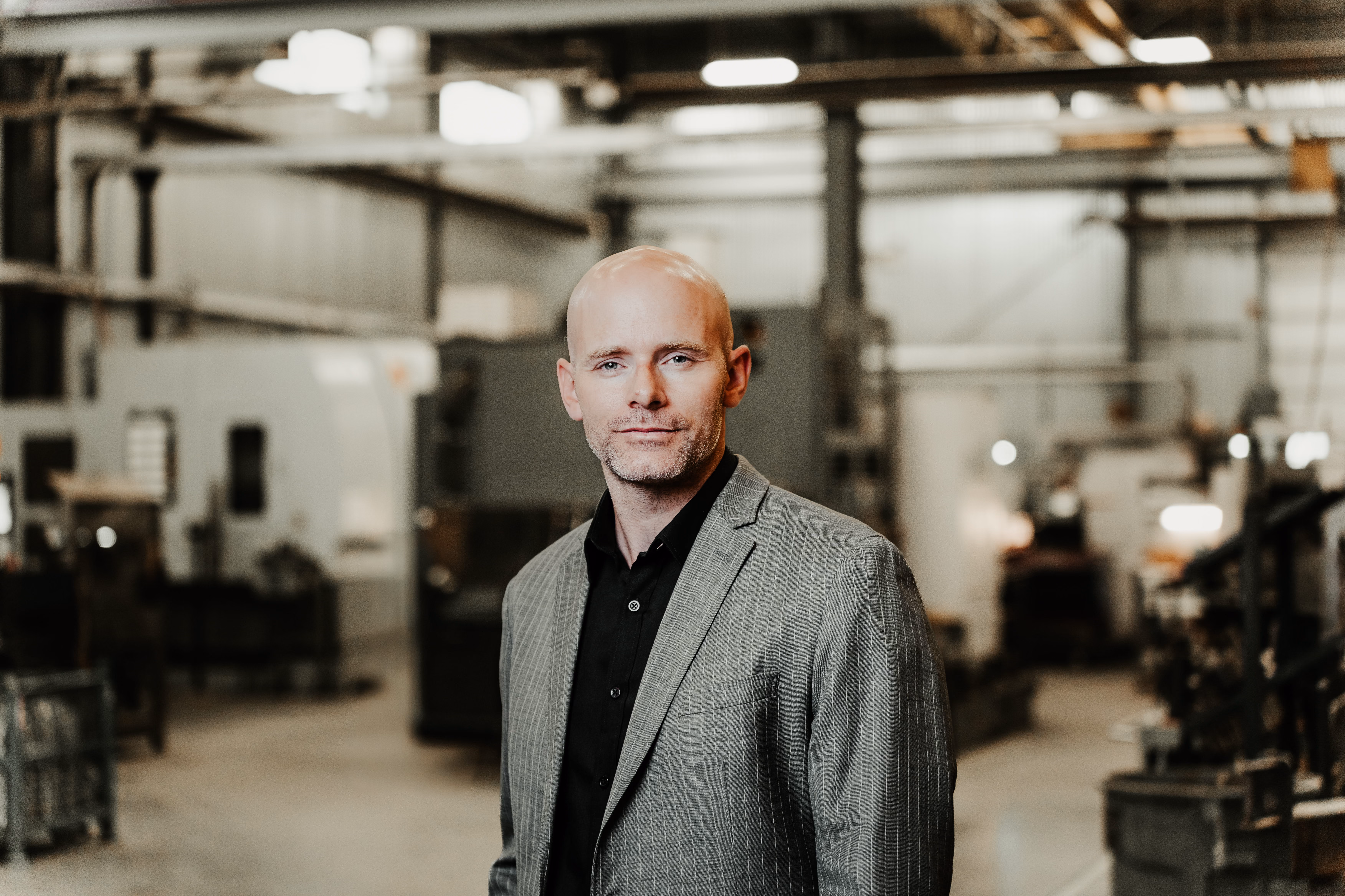 A bald man wearing a gray pinstripe blazer and black shirt stands in an industrial or workshop setting with machinery and equipment blurred in the background.