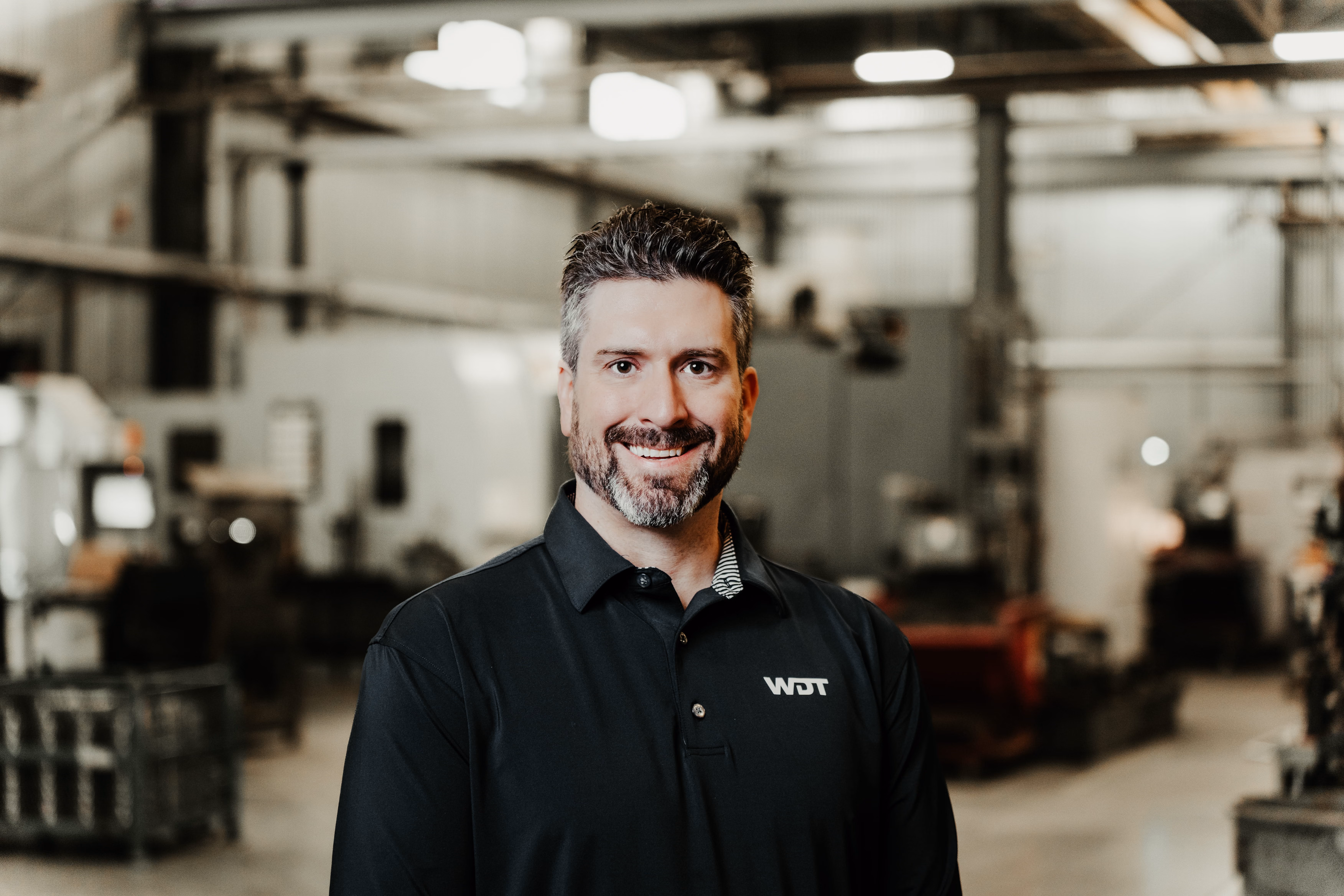 Smiling man with short dark hair and a beard wearing a black WDT polo shirt standing in a blurred industrial workshop background.