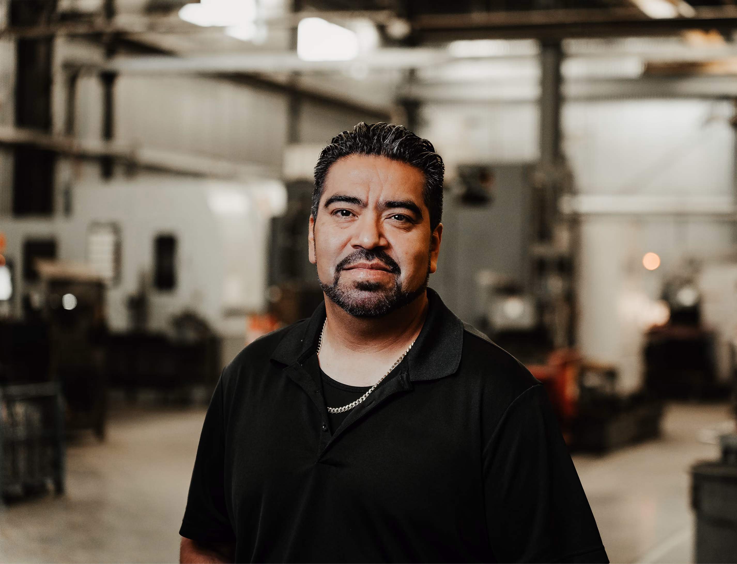 Man with short dark hair and a goatee wearing a black polo shirt and a gold chain standing in an industrial workshop with blurred machinery and equipment in the background.
