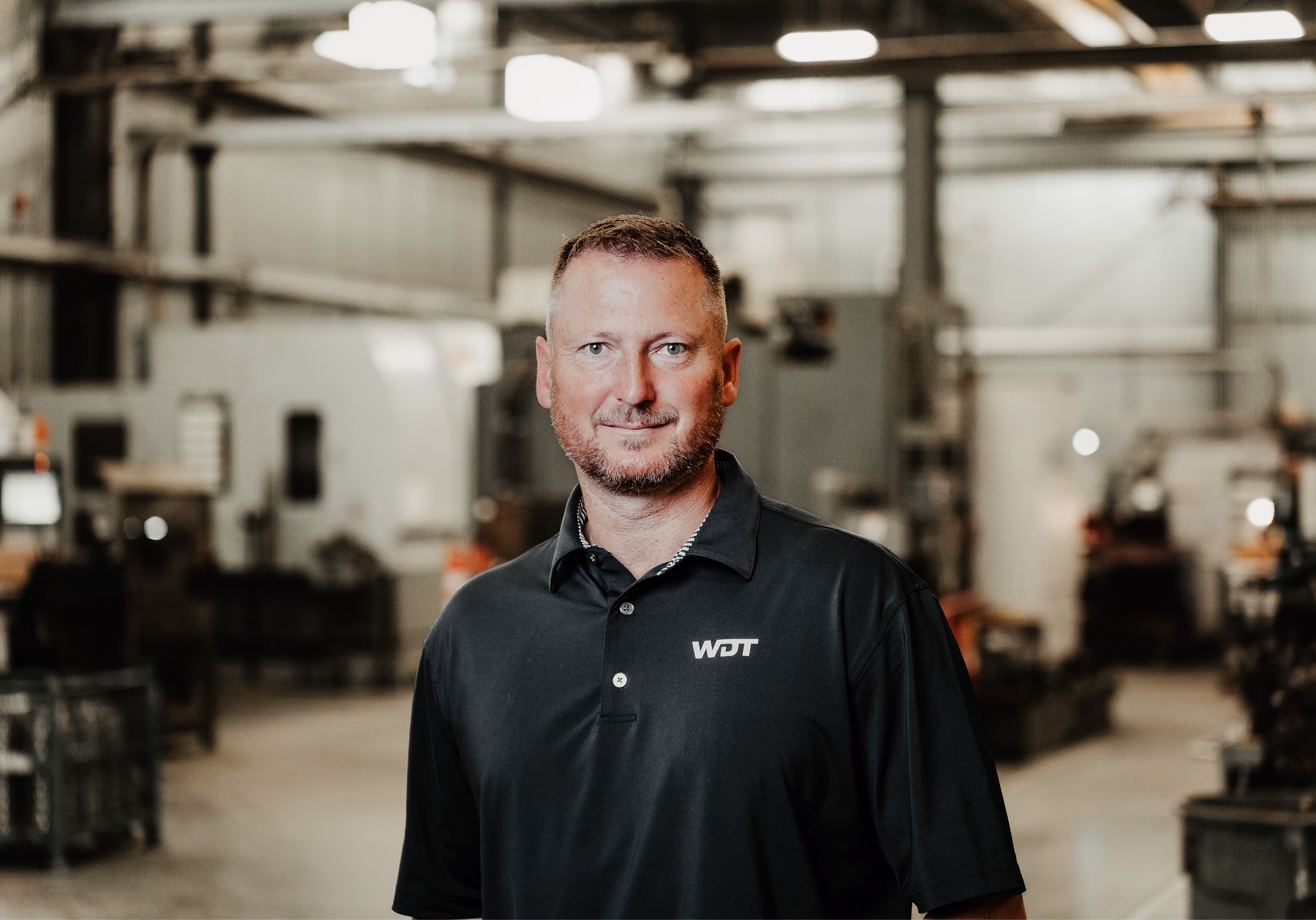 Man with short brown hair and a trimmed beard standing in a spacious industrial workshop wearing a black polo shirt with a WDT logo, with machinery and equipment blurred in the background.